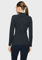 A woman shown from the back wearing a dark charcoal long-sleeve athletic top with a mock neck and tonal lace-up back detail, paired with dark leggings, against a light gray studio background.