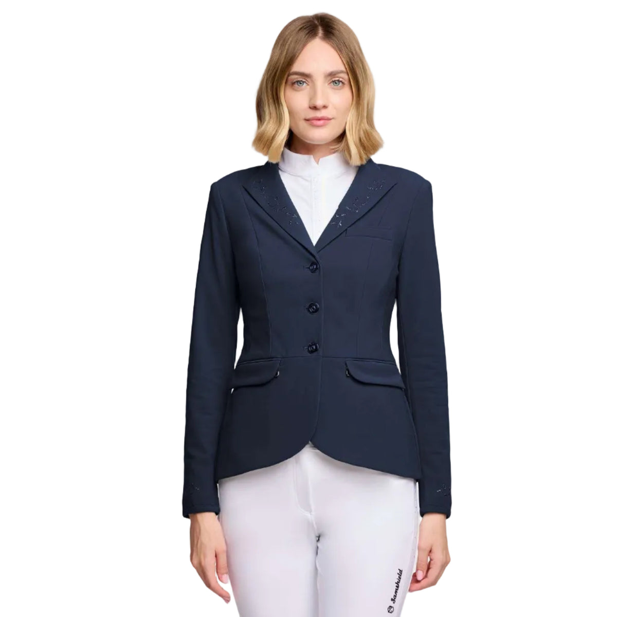 A woman modeling a navy blue equestrian-style blazer with decorative silver studs on the shoulders, worn over a white mock neck top and white riding pants, against a plain white background.