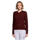 A woman modeling a dark burgundy cable-knit V-neck sweater with a small black embroidered logo on the left chest, paired with white leggings, against a light gray studio background.