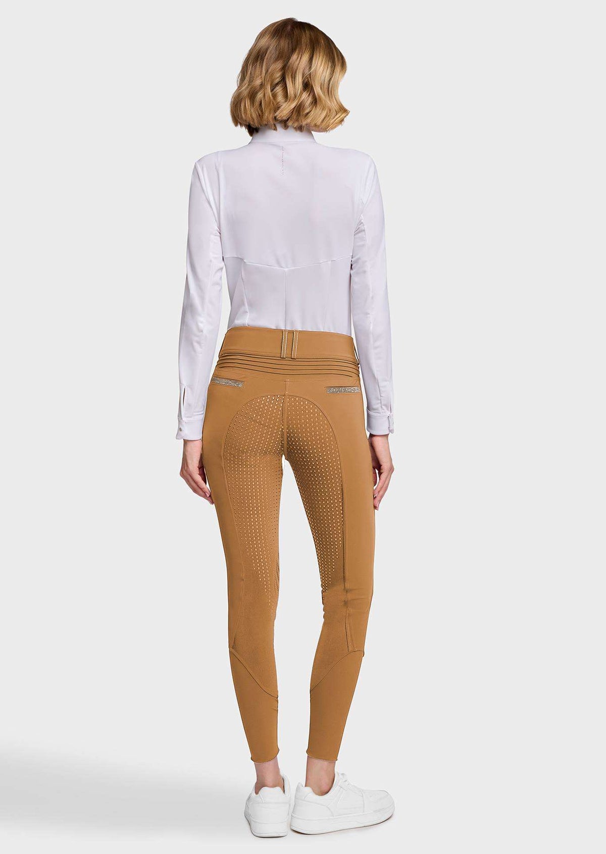 A woman viewed from the back wearing tan biker shorts with a textured grip pattern, a white long-sleeve shirt, and white sneakers, set against a light gray studio background.