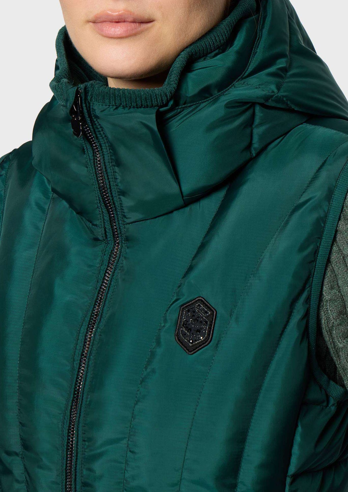 Close-up of a person wearing a dark green hooded puffer vest with a black zipper and a small black shield-shaped logo patch on the chest.