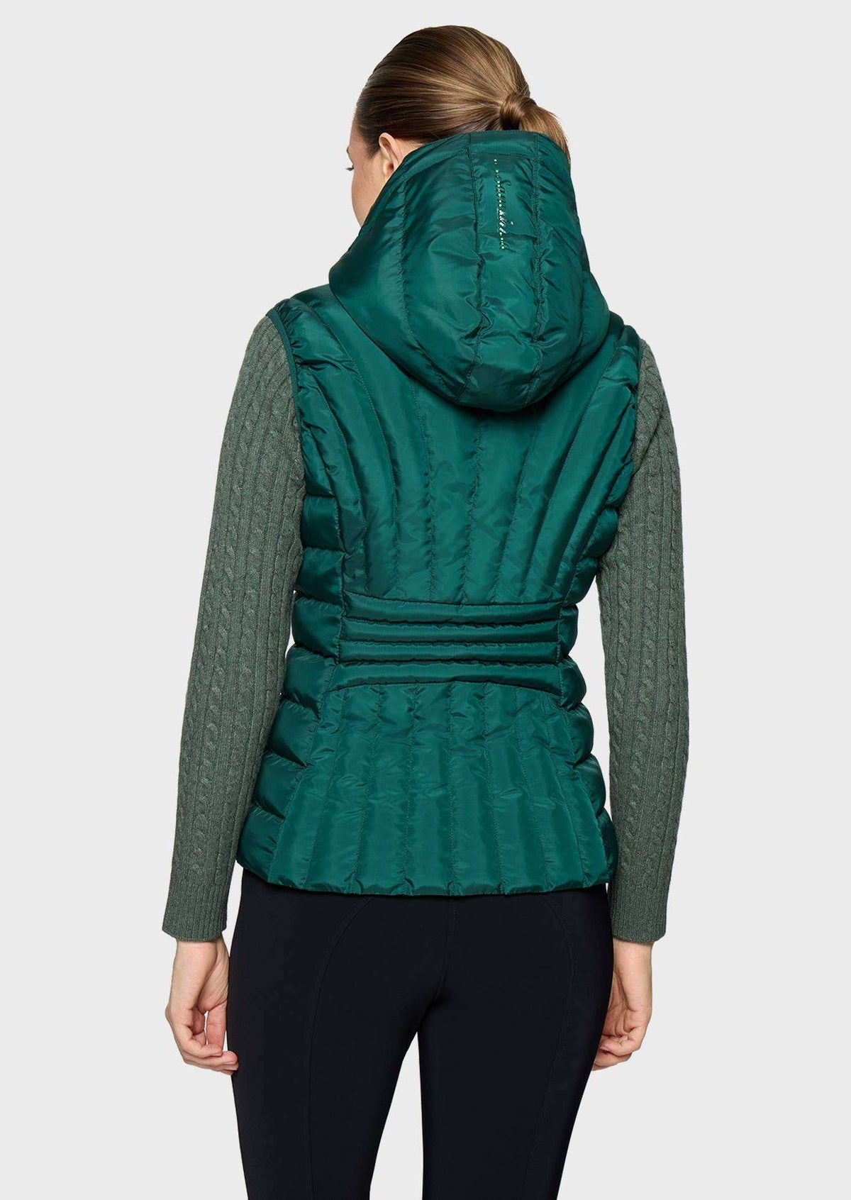 A woman shown from the back wearing a dark teal green hooded puffer vest over a dark green cable-knit long-sleeve top and black leggings, against a light gray studio background.