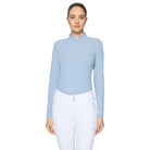 A woman modeling a light blue long-sleeve equestrian quarter-zip top paired with white riding breeches, set against a solid light gray studio background.
