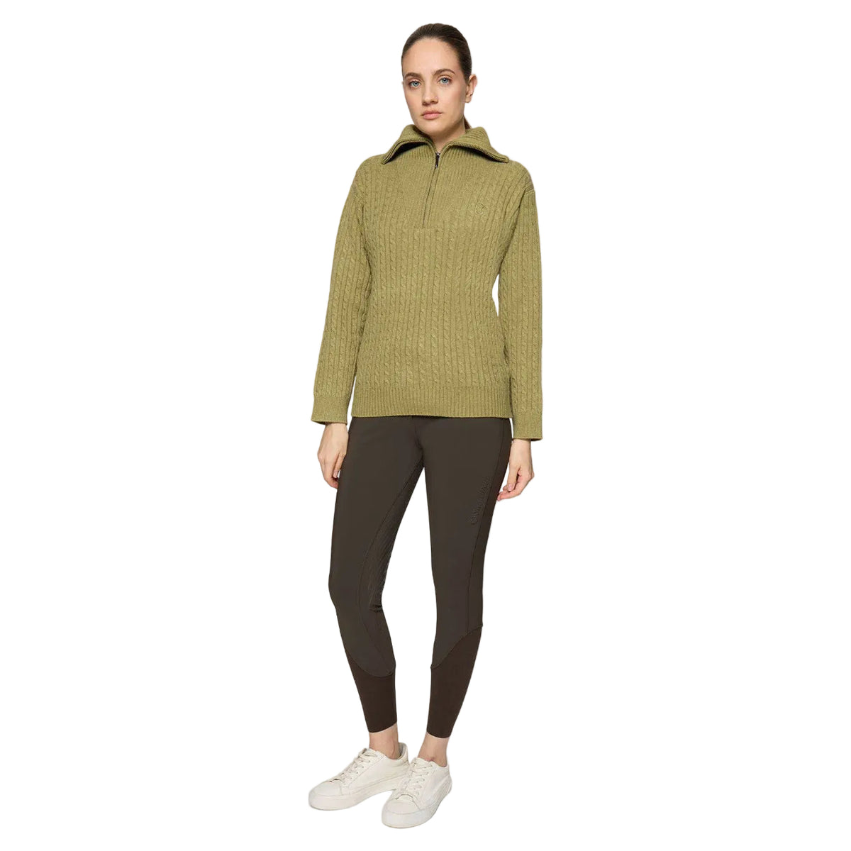 A woman modeling a ribbed olive green half-zip pullover sweater with a stand-up collar, paired with dark brown leggings and white sneakers, against a light gray studio background.