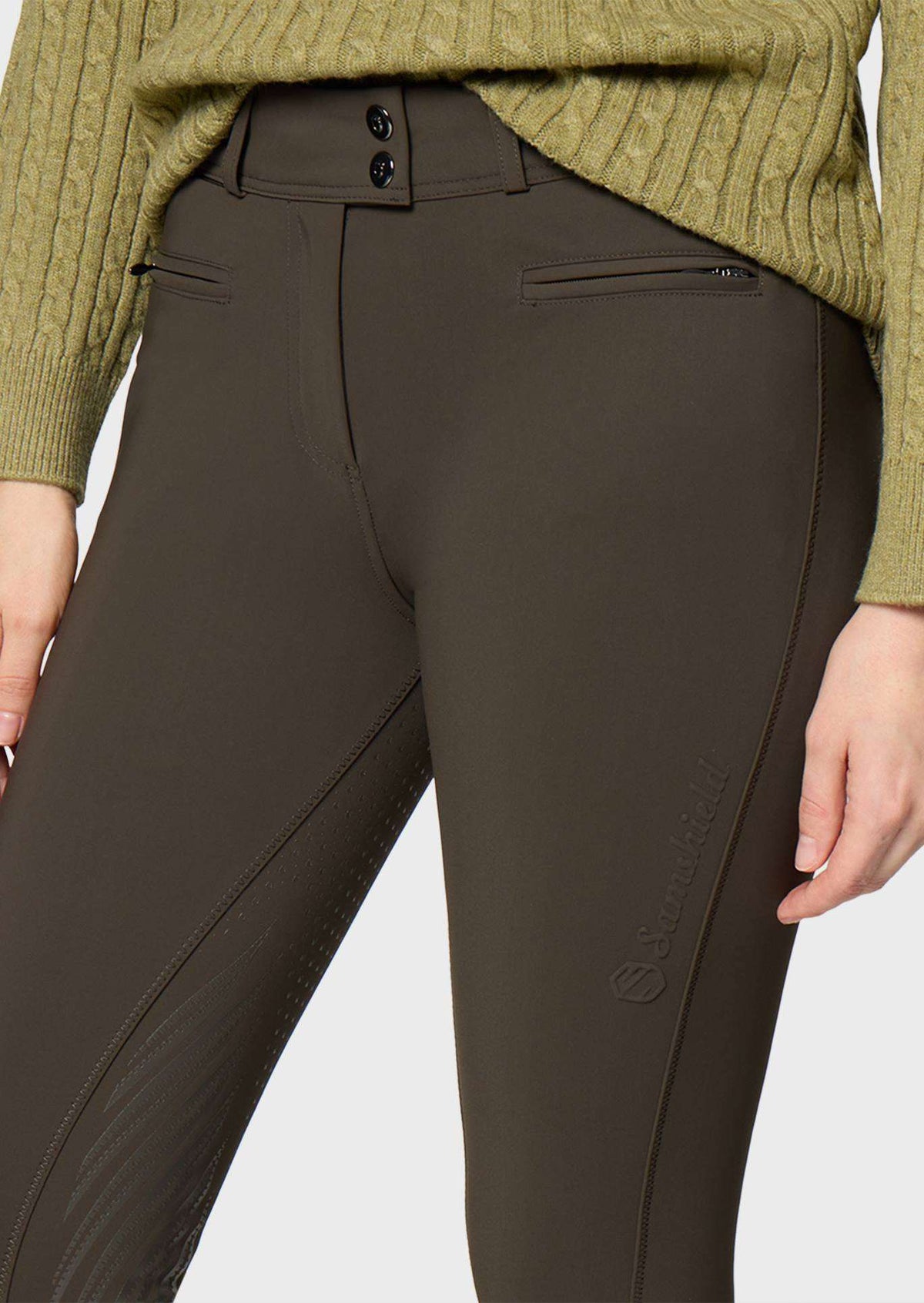 Close-up of a person wearing dark olive riding breeches with a ribbed olive sweater partially visible at the top, against a light gray studio background.