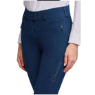 Close-up of a person wearing high-waisted navy blue riding breeches with decorative black embellishments at the waistband, zippered side pockets, and a small gold script logo on the left leg, paired with a white shirt against a light gray background.