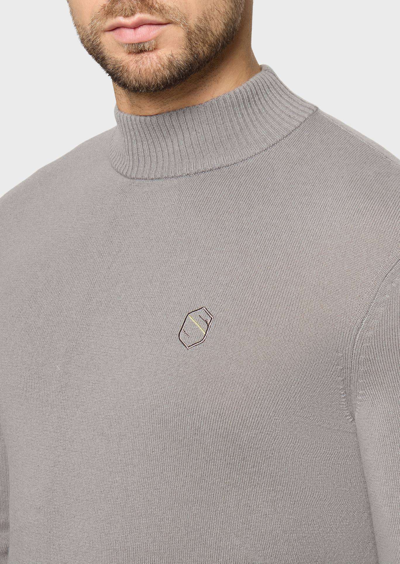 A close-up of a man wearing a light gray ribbed mock neck sweater with a small embroidered geometric logo on the left chest, set against a plain light gray background.