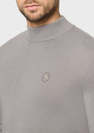 A close-up of a man wearing a light gray ribbed mock neck sweater with a small embroidered geometric logo on the left chest, set against a plain light gray background.