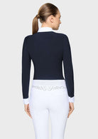 A woman shown from the back wearing a fitted dark navy long-sleeve top with a white collar and white cuffs, paired with white pants featuring a decorative embroidered waistband.