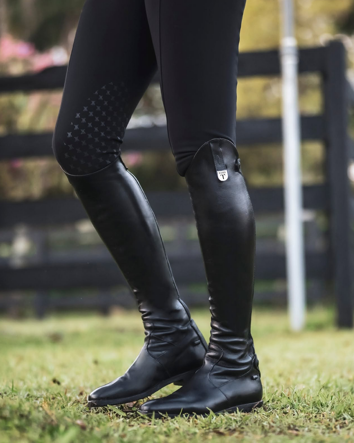 Black knee-high riding boots worn with black riding tights on a person standing on grass in front of a blurred fence.