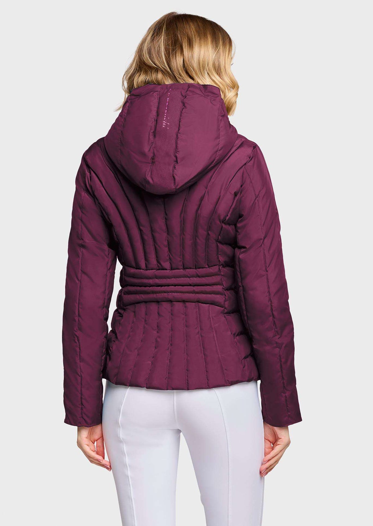 A woman viewed from the back wearing a quilted hooded jacket in deep plum purple with a matching tie belt, paired with white pants, against a light gray studio background.