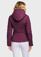 A woman viewed from the back wearing a quilted hooded jacket in deep plum purple with a matching tie belt, paired with white pants, against a light gray studio background.