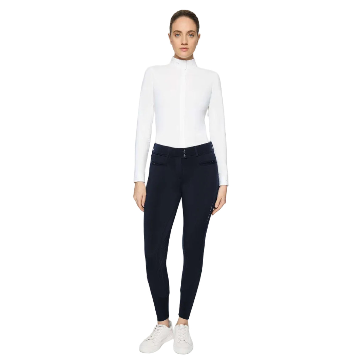 A woman modeling a white long-sleeve mock neck top and dark navy fitted pants with white sneakers, photographed against a light gray studio background.