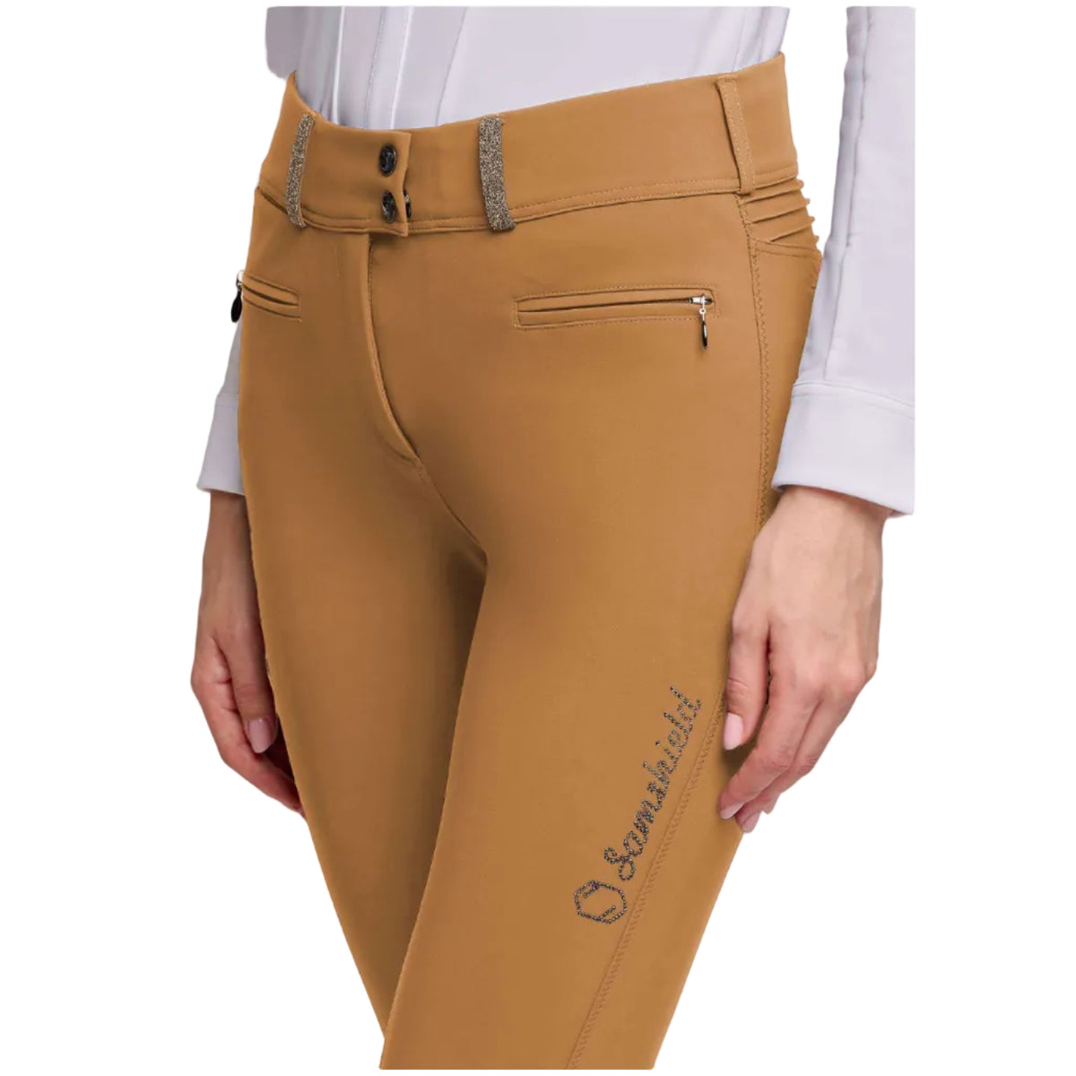 A close-up of a model wearing brown equestrian riding breeches with a decorative waistband, zippered side pockets, and a black embroidered logo on the left leg, paired with a white shirt.