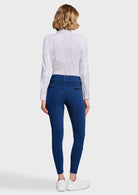 A woman viewed from the back wearing a white long-sleeve shirt and blue patterned riding breeches with white sneakers, against a light gray studio background.
