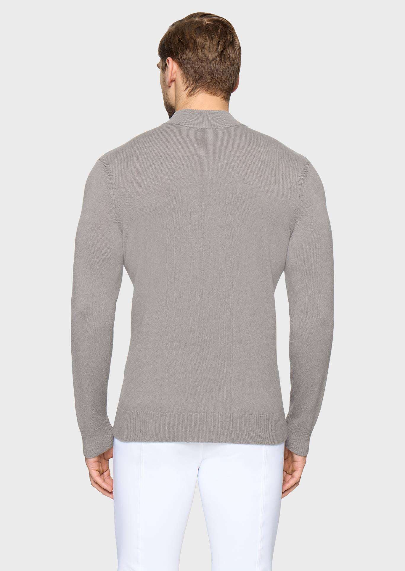 A man viewed from the back wearing a light gray ribbed knit sweater and white trousers, standing against a plain light gray studio background.