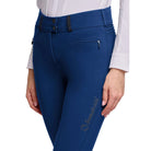 Close-up of a person wearing blue riding breeches with a white shirt. The pants have a decorative waistband, zippered side pockets, and a small gold embroidered 'Somerset' logo on the lower leg.