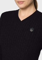 Close-up of a woman wearing a navy blue cable-knit V-neck sweater with a small circular embroidered logo on the chest, against a light gray background.