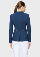 A woman shown from the back wearing a navy blue tailored blazer with a peplum-like waist and small embellished detail at the center back, paired with white pants, against a light gray studio background.