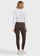 A woman shown from the back wearing dark brown riding breeches, a white long-sleeve shirt, and white sneakers against a light gray studio background.