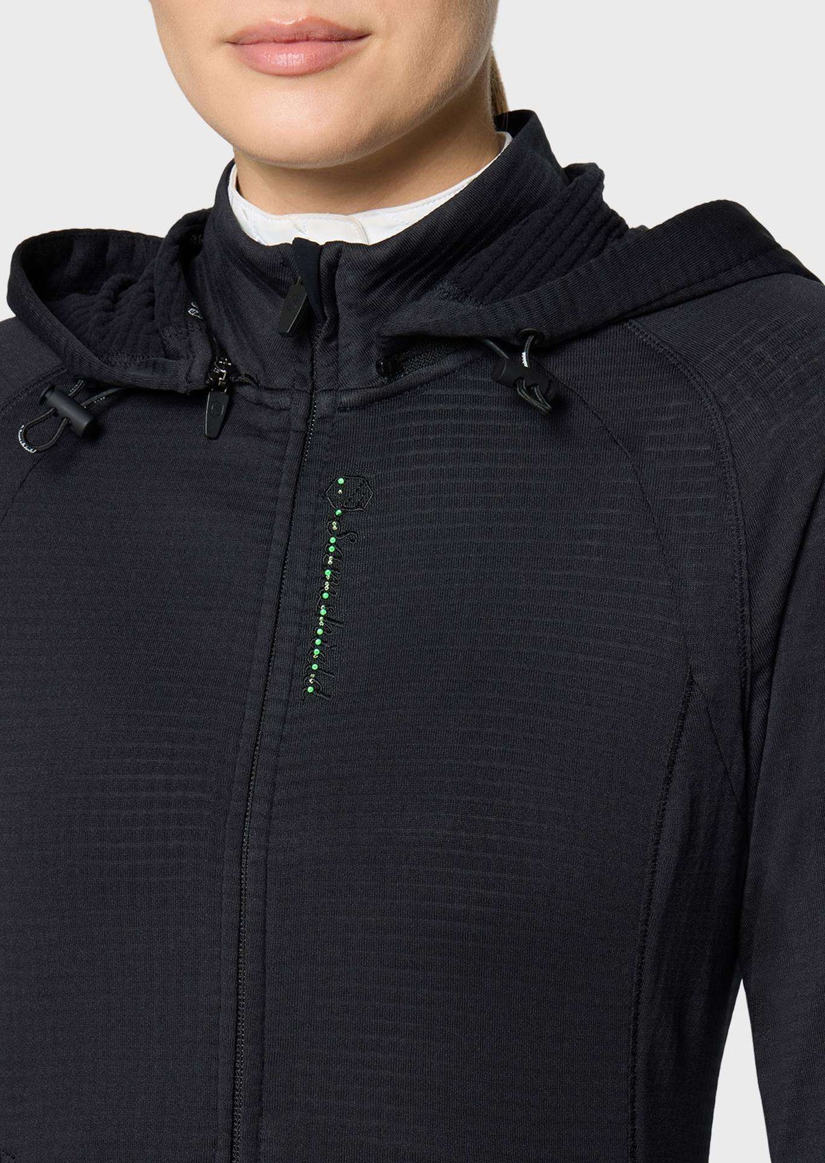 Close-up of a person wearing a black hooded zip-front jacket with a subtle textured knit fabric and a small green vertical accent near the chest, photographed against a light gray background.