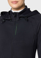 Close-up of a person wearing a black hooded zip-front jacket with a subtle textured knit fabric and a small green vertical accent near the chest, photographed against a light gray background.