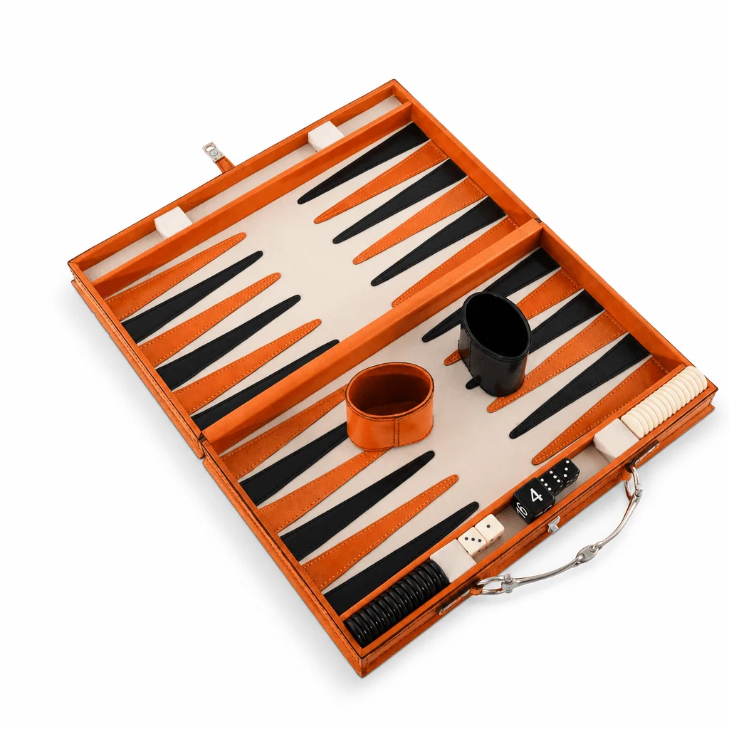 An open backgammon set in a bright orange leather case, shown on a clean white background. The game board has alternating orange and black points on a cream-colored surface, with matching orange and black cups, dice, and a comb inside the case.