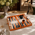 An orange leather backgammon set with black and white checkers and cups, displayed on a beige tablecloth in a styled dining room setting.