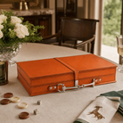 An orange-brown leather checkers box with silver metal hardware sits on a beige tablecloth in a dining room setting, surrounded by checkers, dice, a folded napkin with a horse embroidery, and a glass of white wine.