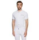 A man modeling a white short-sleeve polo shirt with a white button-down collar and a small black embroidered logo on the left chest, paired with white trousers, against a light gray studio background.