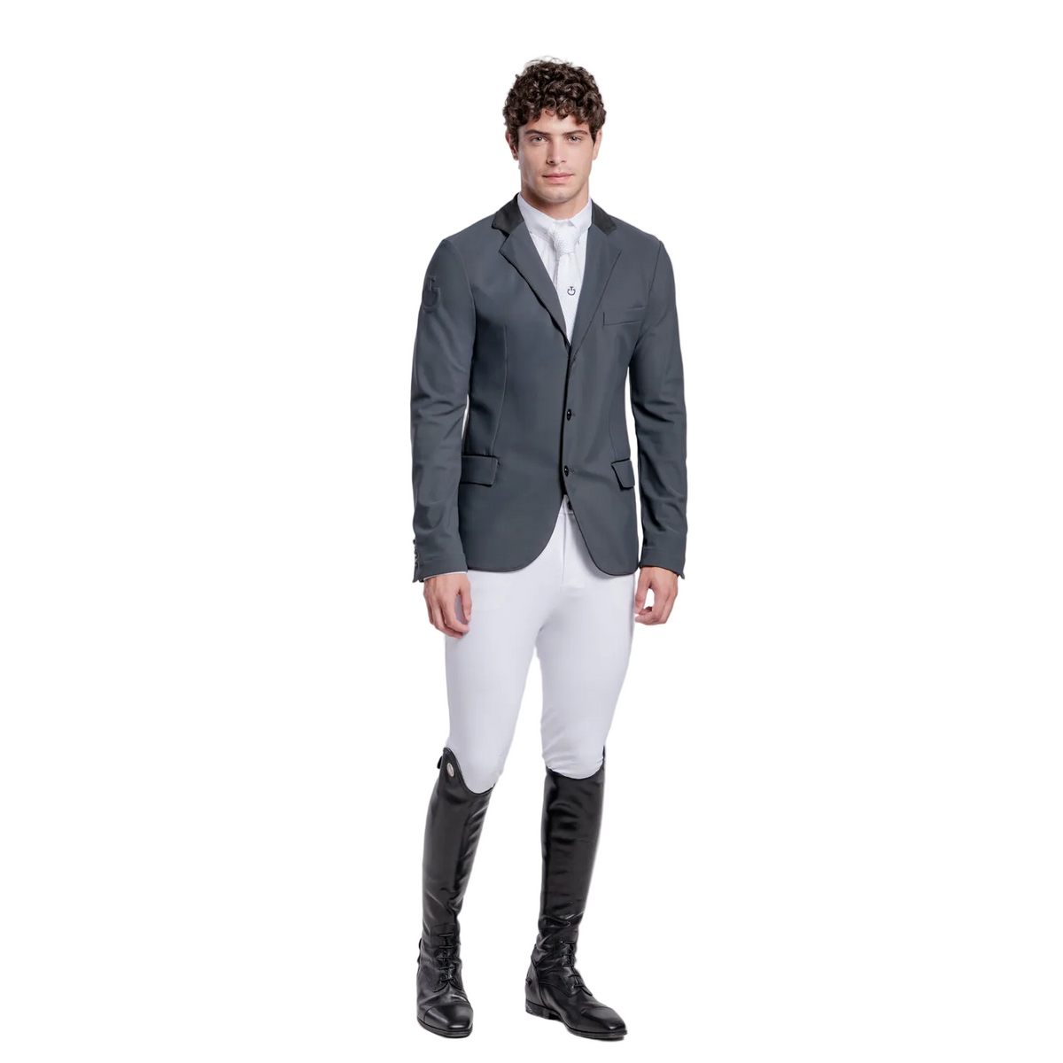 A man modeling a dark gray equestrian sport coat, white breeches, and black riding boots against a light gray studio background.