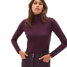A woman modeling a dark plum long-sleeve mock neck top with  'DADA' printed on the lower sleeve, paired with equestrian riding pants, against a solid white background.