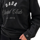 A close-up of a person wearing a black sweatshirt with white printed text reading 'DADA Cheval Club est. 2012' on the front.