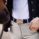 Person wearing a dark jacket, light shirt, beige pants, and a gold belt buckle with a horse in the foreground.