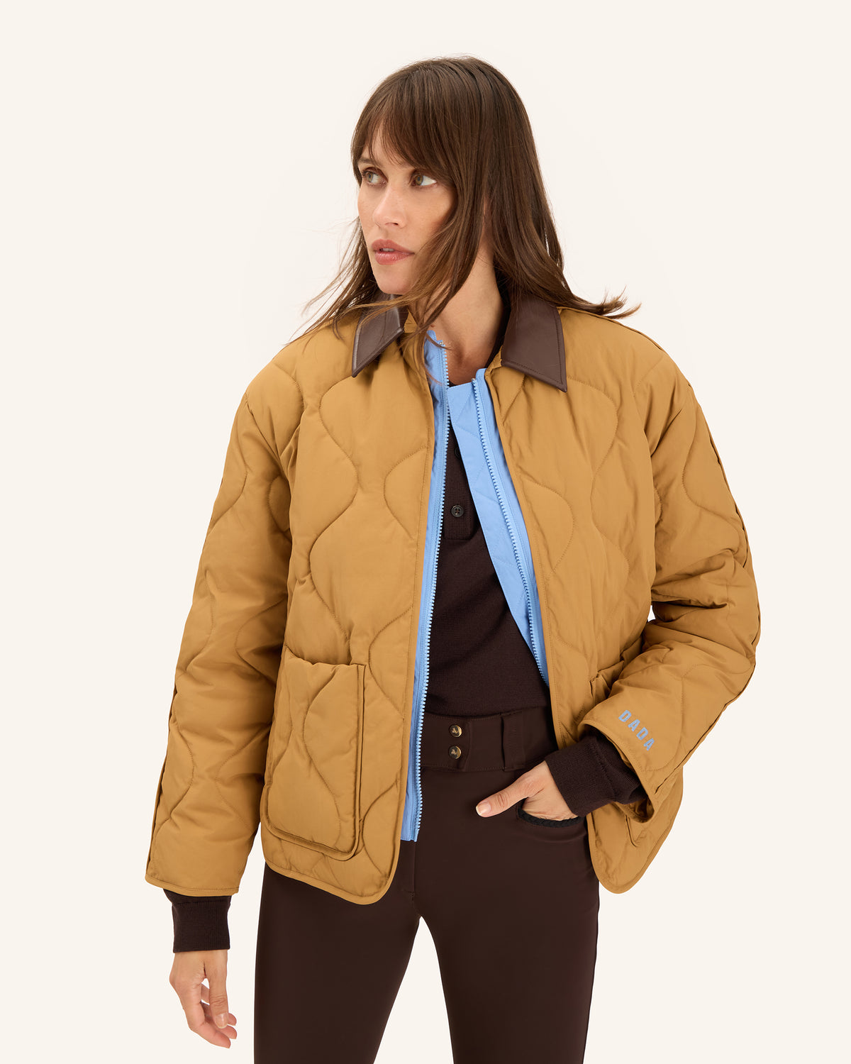 A woman modeling a quilted tan jacket with dark brown collar and cuffs, worn open over a light blue shirt and dark brown pants, against a plain off-white background.