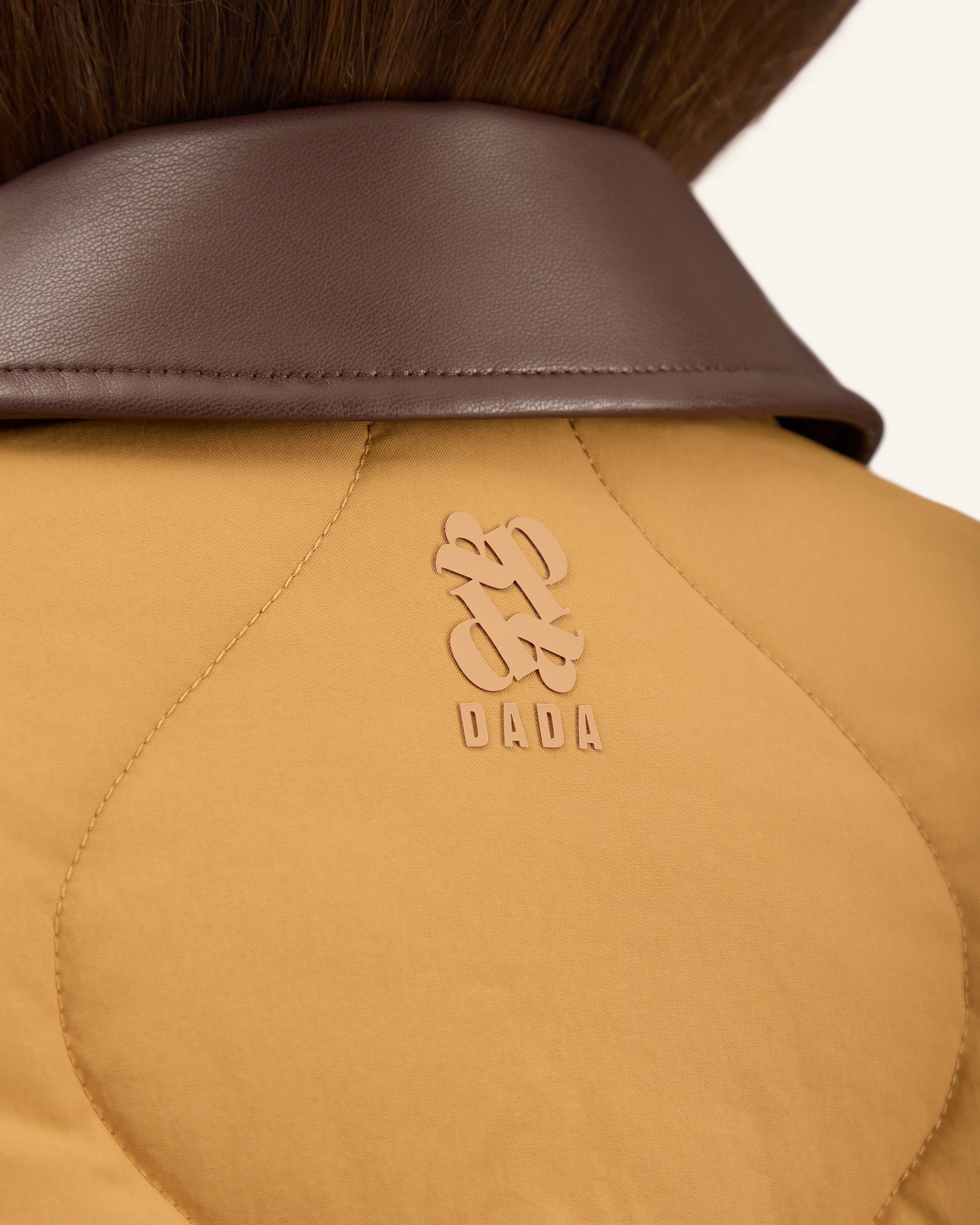 Close-up of the back of a mustard yellow quilted puffer jacket with a brown collar, featuring an embossed DADA logo on the upper back.