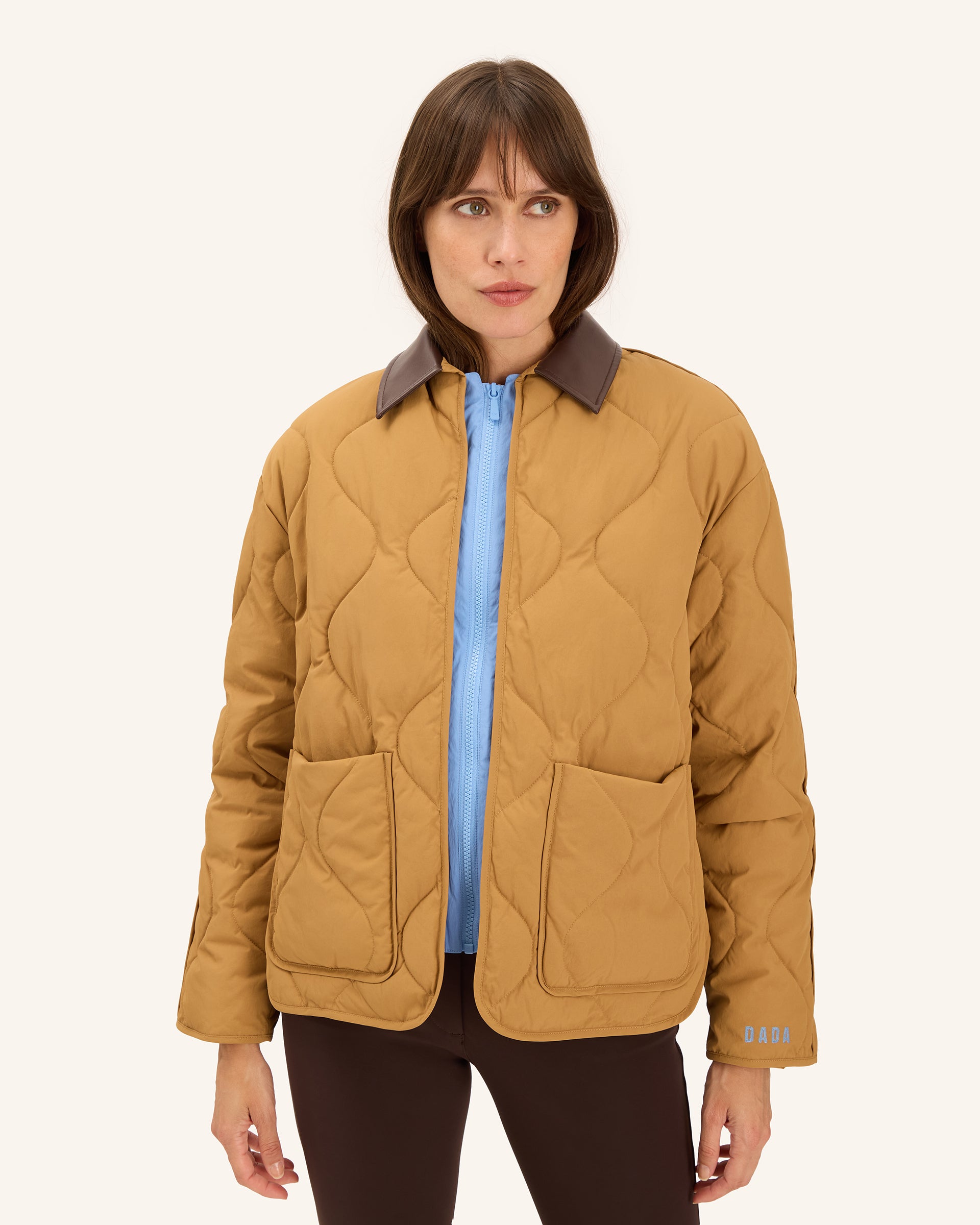 A woman wearing a quilted mustard yellow jacket with a brown collar, layered over a light blue shirt and dark brown pants, against a solid off-white background.
