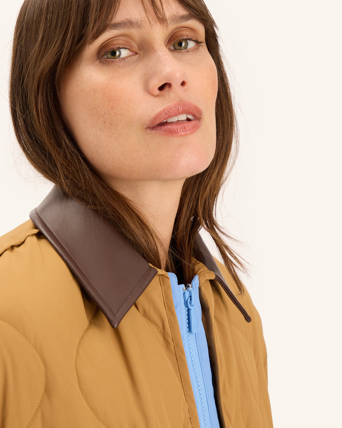Close-up of a woman wearing a tan quilted jacket with a contrasting dark brown collar and a light blue zipper.