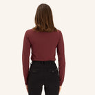 A woman is shown from the back wearing a fitted burgundy long-sleeve top, black fitted pants, and black knee-high boots against a plain off-white background.