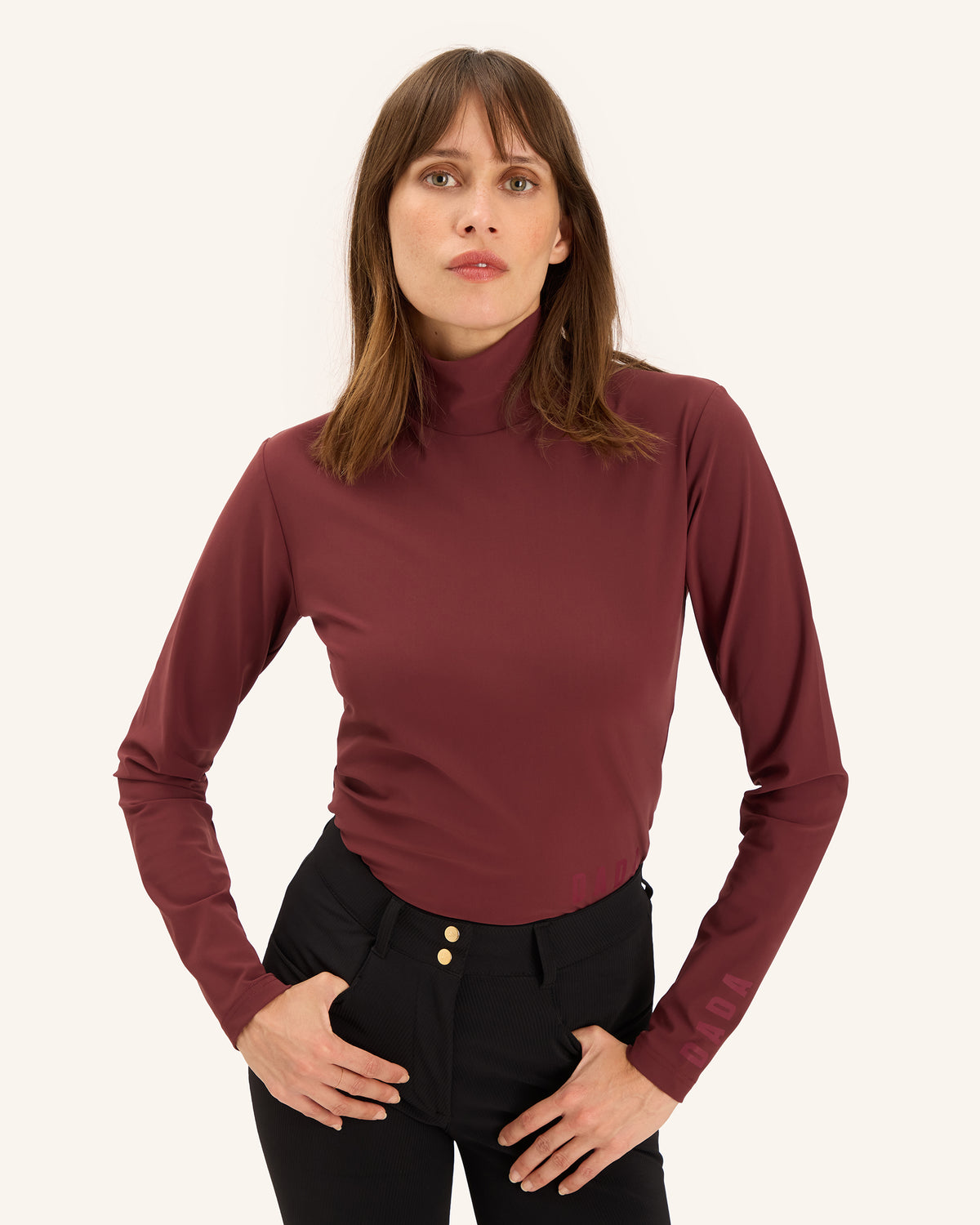 A woman modeling a fitted, long-sleeve mock neck top in a deep burgundy color, paired with black pants, against a solid off-white background.