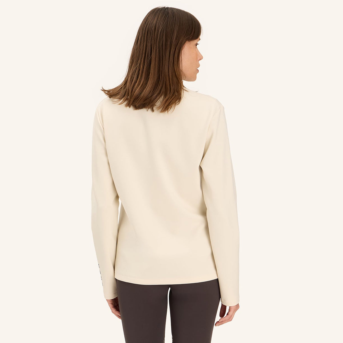 A woman is shown from the back wearing a cream-colored knit sweater, dark gray skinny jeans, and black knee-high riding boots against a solid off-white background.