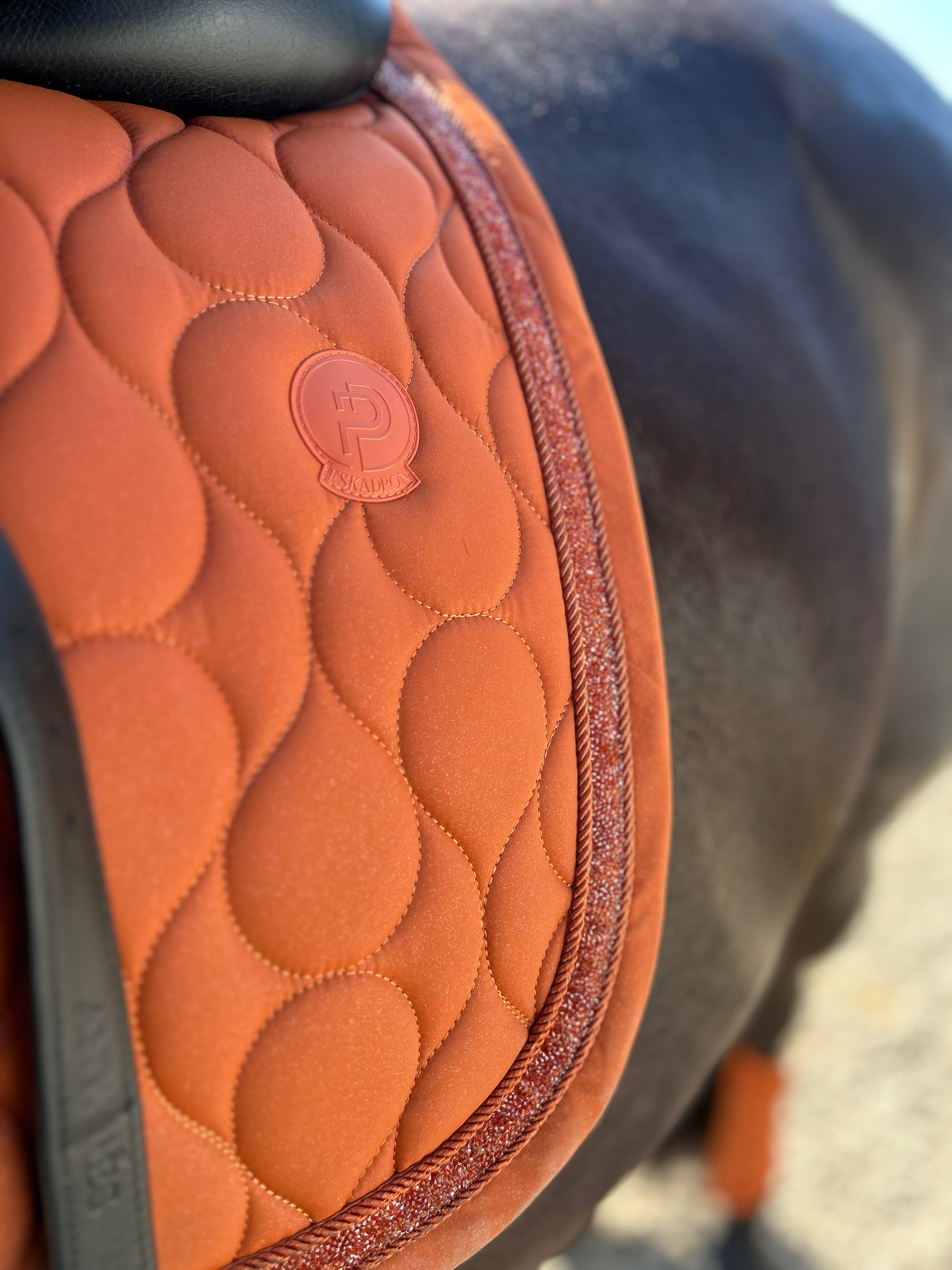 Close-up of an orange quilted horse saddle pad with a scalloped pattern and a circular logo patch, shown on a horse.
