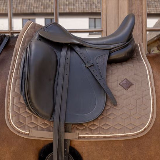 A black leather horse saddle resting on a brown horse, positioned over a tan quilted saddle pad with cream piping.