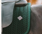 A close-up of an equestrian saddle pad in forest green with a chevron quilted pattern and a silver diamond-shaped logo patch, shown on a horse.