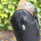 A close-up of a black leather boot with a silver oval logo badge, shown outdoors with green foliage in the background.