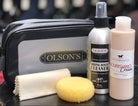 Olson's leather care kit including a spray boot cleaner bottle, cream, and sponge on a table with shoes in the background.