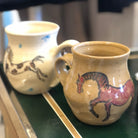 Two ceramic mugs with horse designs on a green surface.