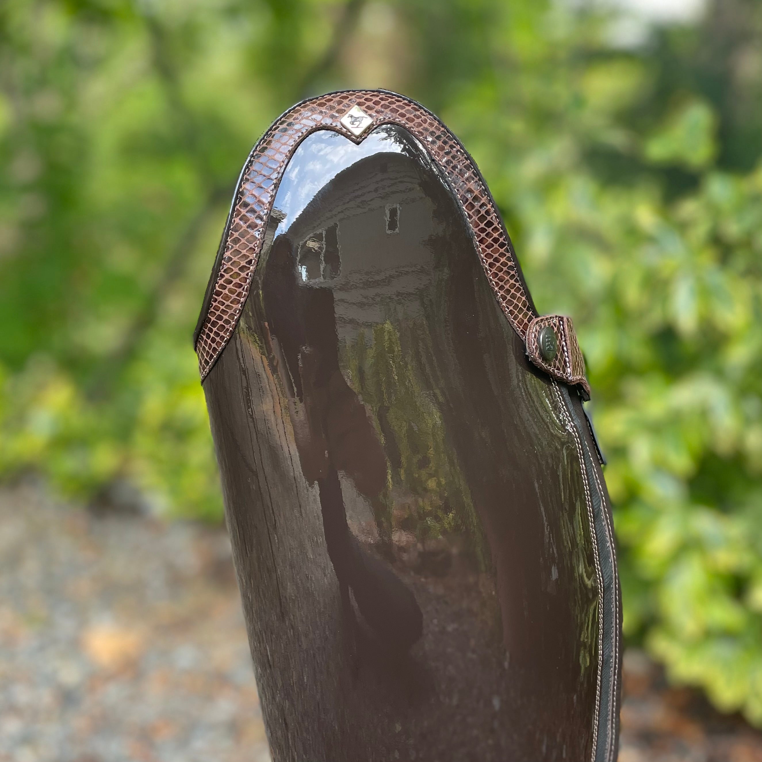A close-up of a glossy dark brown rain boot with textured brown trim, photographed outdoors against a blurred green foliage background.