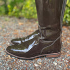 A pair of glossy dark brown patent leather riding boots with a low block heel, shown on a gravel path with a blurred green garden background.