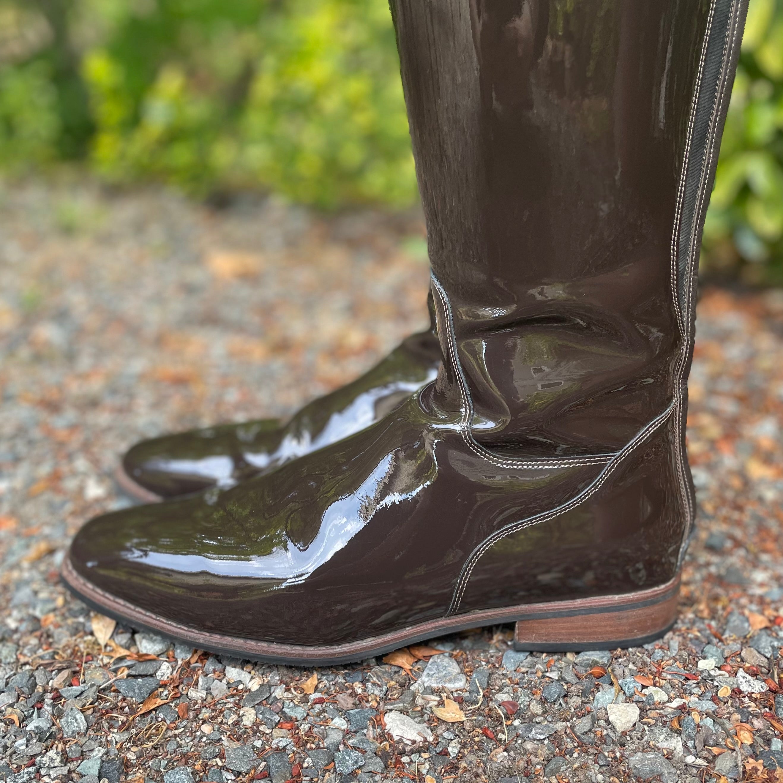 A pair of glossy dark brown patent leather riding boots with a low block heel, shown on a gravel path with a blurred green garden background.